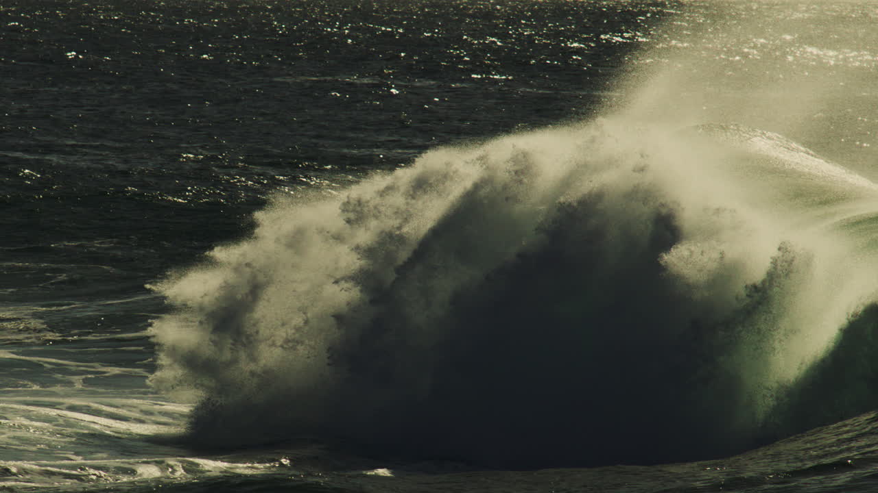 Low light catches on peeling wave lip as spray and mist roll across sea in slow motion crash