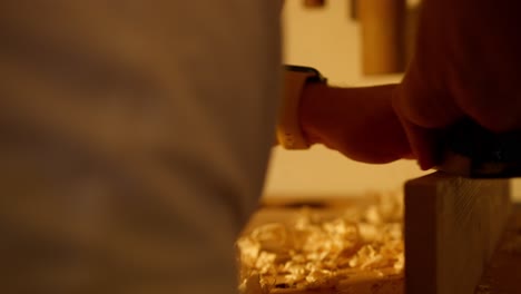 Close-up-of-a-man-sanding-a-wooden-board-while-pieces-of-it-jump-off-in-workshop