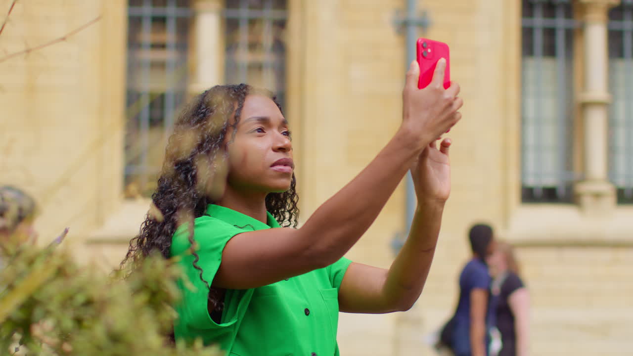 영국 옥스퍼드에서 휴가를 보내는 여성 관광객이 브로드 스트리트를 따라 걸어서 휴대전화로 사진을 찍고 있습니다.