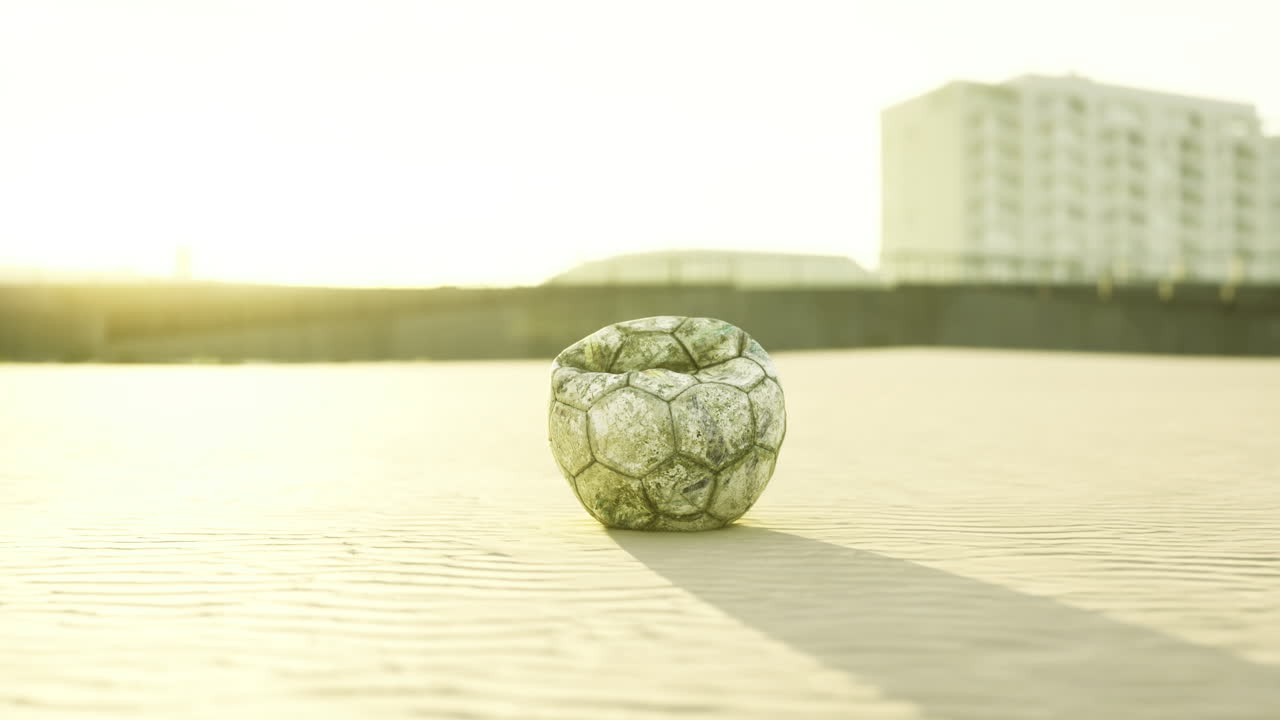 Old soccer ball resting on sandy beach during sunset near urban background