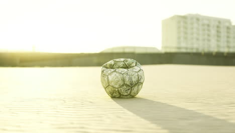 old soccer ball resting on sandy beach during sunset near urban background