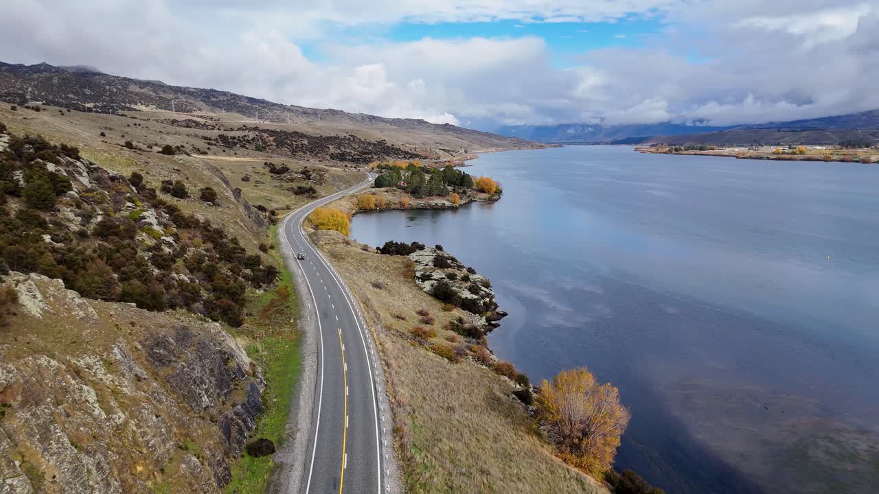 A drone smoothly flies above a winding lakeside road with a car driving, capturing reflections on calm water and scenic rural landscape in daylight