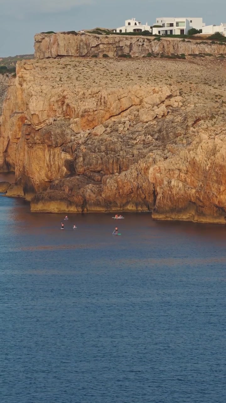 Vista aerea verticale di canoe sotto scogliere calcaree con case costiere sopra, Cala Morell, Minorca.