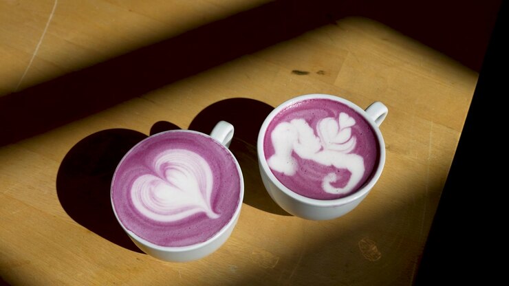 Two purple ube lattes display white foam artwork, heart and complex white pattern, in ceramic cups on a wooden counter