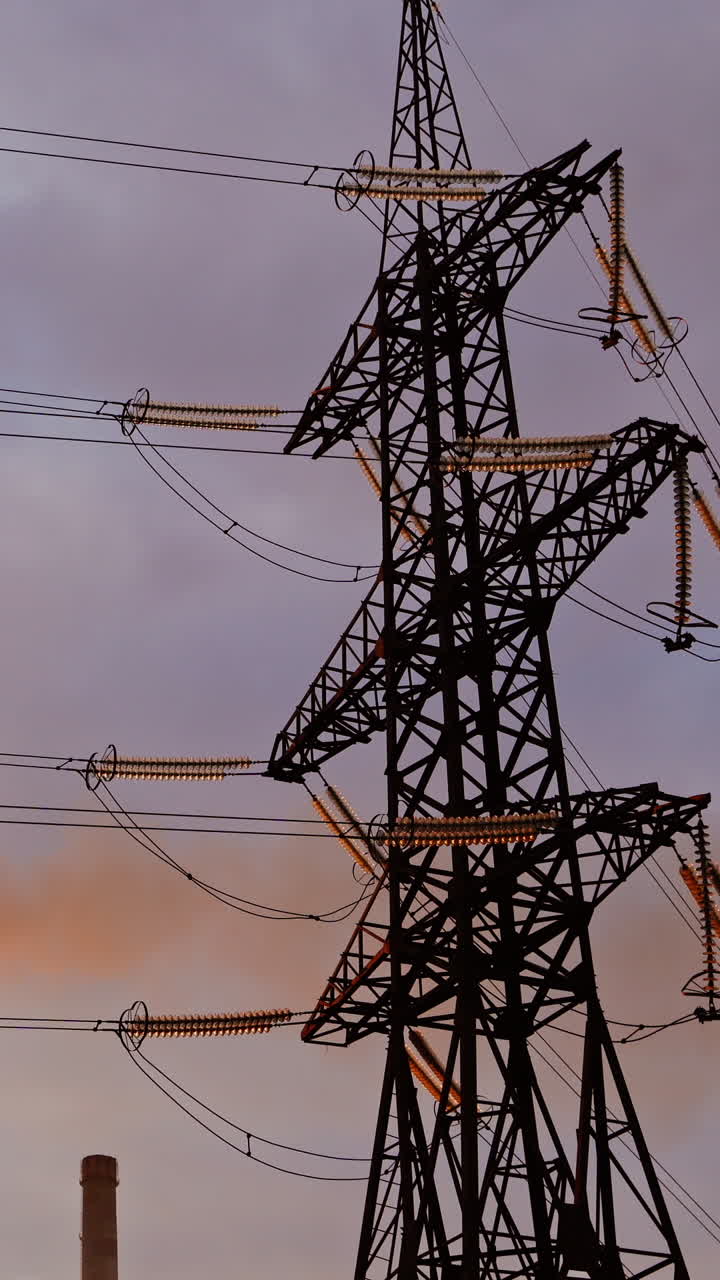 Electricity pylon on industrial chimney background. High-voltage tower and pipe of power plant with smoke on evening sky. Harmful smoke polluting the environment. Vertical video