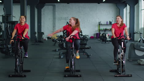 grupo de amigas sonrientes mujeres clase de ejercicio, entrenamiento, girar en bicicleta estacionaria en un gimnasio moderno