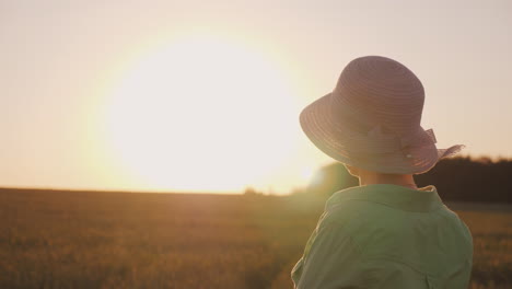 a female farmer in a hat looks at the horizon above a field of wheat enjoying the sunset the back vi