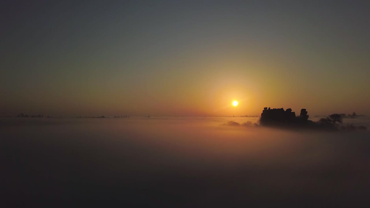 Drone flies above low lying clouds with nothing but a large mound of rocks peaking above to capture the first rays of sunlight