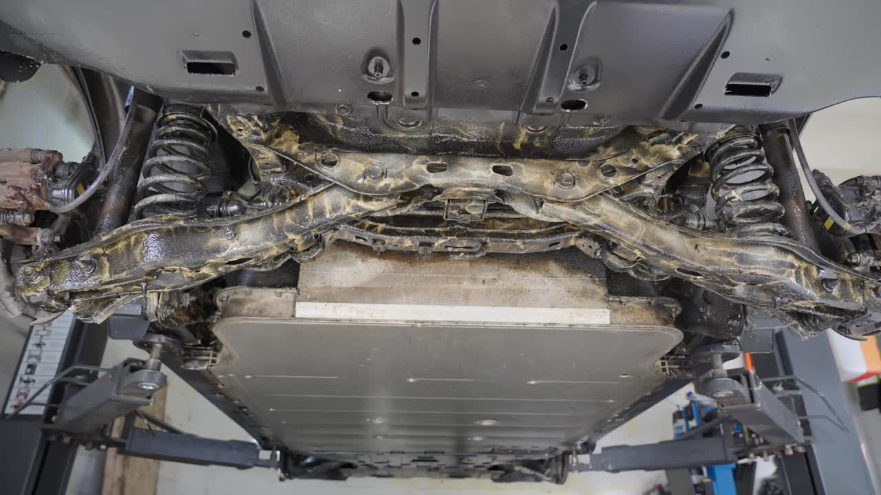 Rear undercarriage of electric car after being sprayed with lanolin rustproofing product, crossmember, control arms, sway bar, battery compartment visible, left to right slider shot