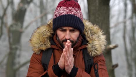 bearded caucasian male standing in forest and warming up his hands with breath during cold winter day, slow motion