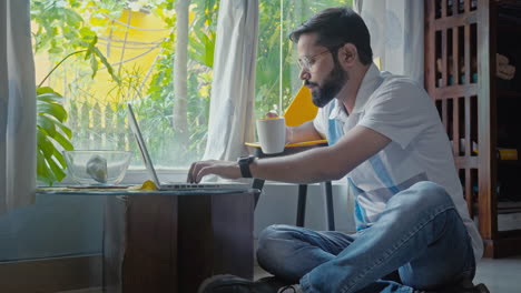 man working on laptop while drinking coffee at home