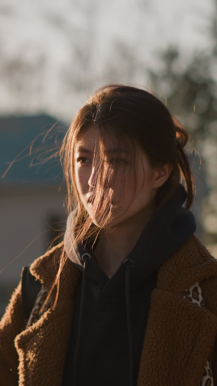 una chica con un abrigo marrón y una sudadera con capucha está caminando débilmente, con una expresión de cansancio y contemplación. su cabello está suavemente arruinado por el viento, momento de reflexión tranquila en un entorno urbano