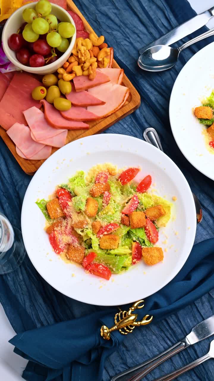 Insalata Caesar fresca con pomodori e crostini vicino a un vassoio di carne. Vista dall'alto verticale della ciotola dell'insalata e del tagliere di stuzzichini su tessuto blu spiegazzato