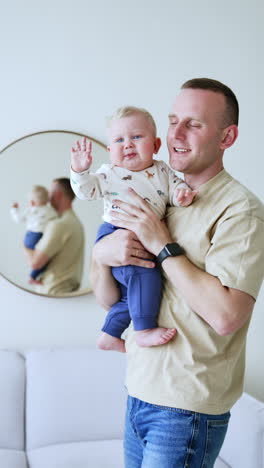 father is holding his cute baby boy standing at the mirror. infant looks aside waving his hand. vertical video.