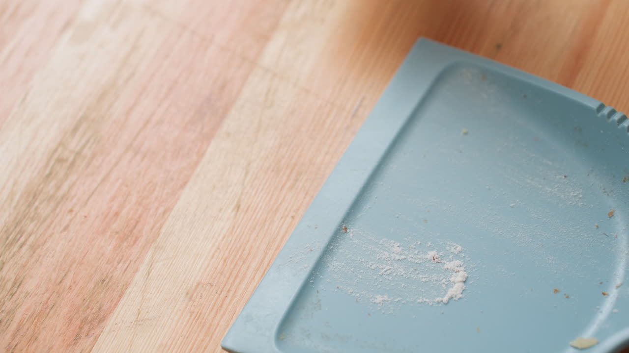 Close up of person sweeping flour and crumbs into blue dustpan with brush on wooden table, showing cleaning process, household tidying, and focus on hygiene during daily indoor routine