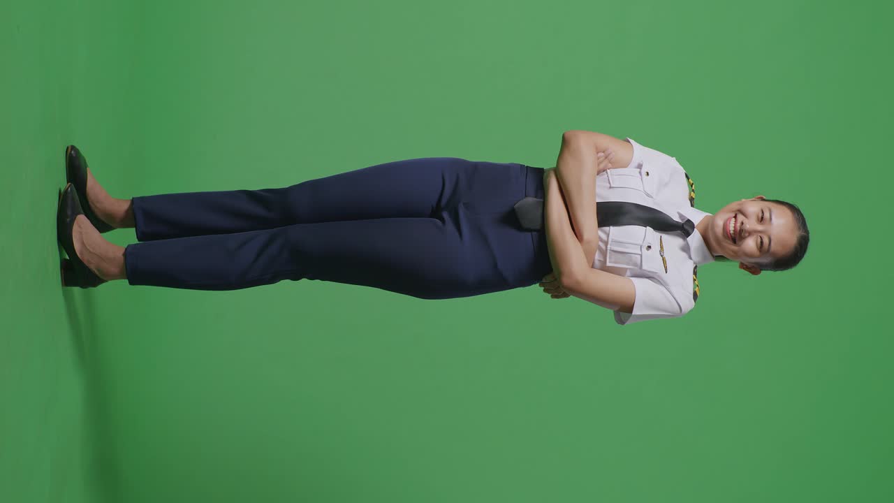 으로 보이는 아시아 여성 조종사가 미소 짓고 녹색 스크린 배경 스튜디오에 서 있는 동안 카메라에 팔을 고 있습니다.