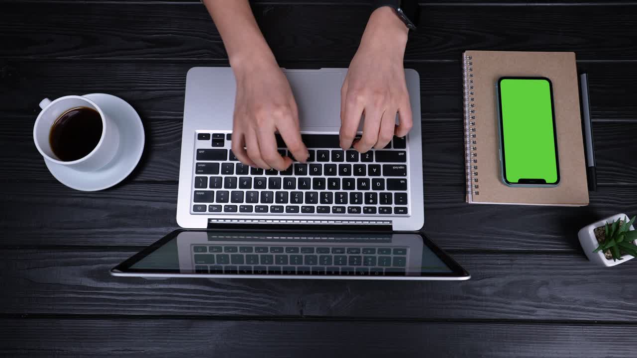 Top view of the hands of a girl who type, writes emails on an laptop. Near the laptop is a smartphone with a green screen and a chrome key. Place for your advertising content. Slow motion. Close up