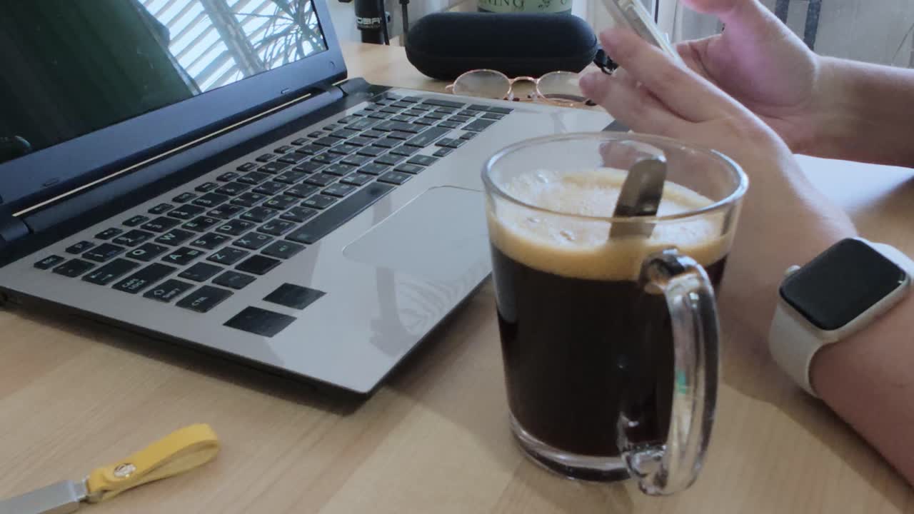Person taking a casual break, browsing on their smartphone at a desk with laptop and coffee.