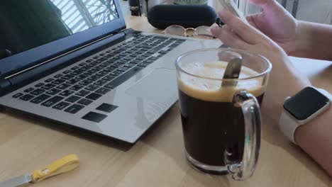 person taking a casual break, browsing on their smartphone at a desk with laptop and coffee.
