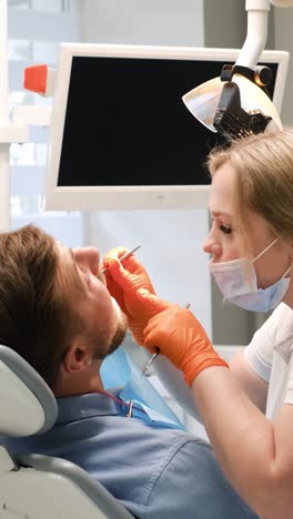 a man in a dental chair is having his teeth examined by a dentist, with his hands and arms at rest while the dentist checks his mouth and gestures for him to open wide