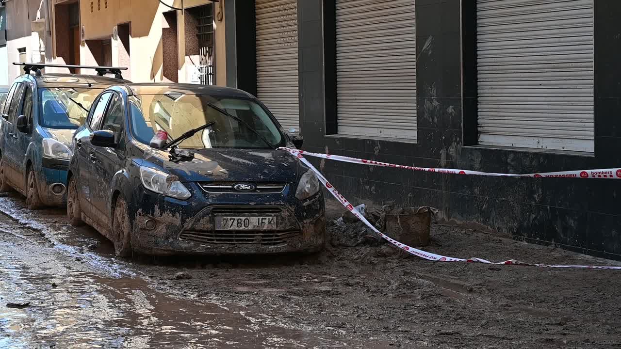 Cars parked on a flooded urban street, covered with mud, indicating flood damage in Valencia. The scene reflects climate change impacts