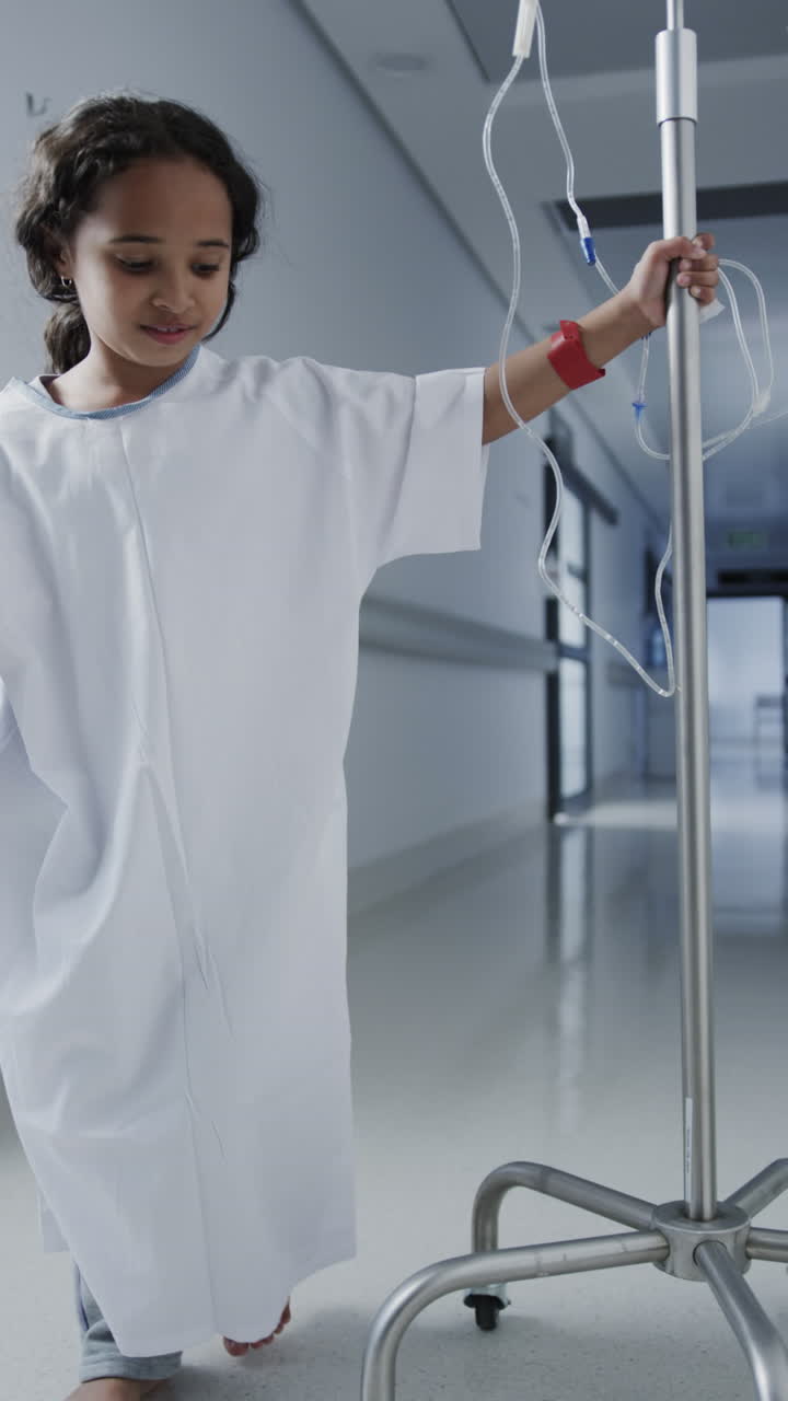 Vertical video of portrait of happy biracial girl walking with drip in hospital in slow motion