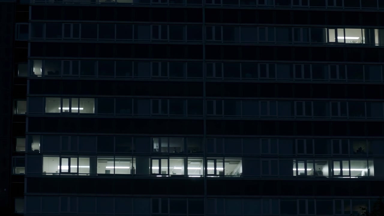 Una vista laterale di un moderno edificio urbano caratterizzato da finestre in vetro e una struttura in cemento, ambientato in un'atmosfera neutra con toni blu scuro e neri, per uso architettonico ed editoriale
