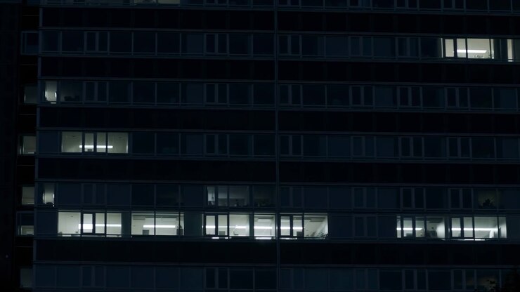 Una vista laterale di un moderno edificio urbano caratterizzato da finestre in vetro e una struttura in cemento, ambientato in un'atmosfera neutra con toni blu scuro e neri, per uso architettonico ed editoriale