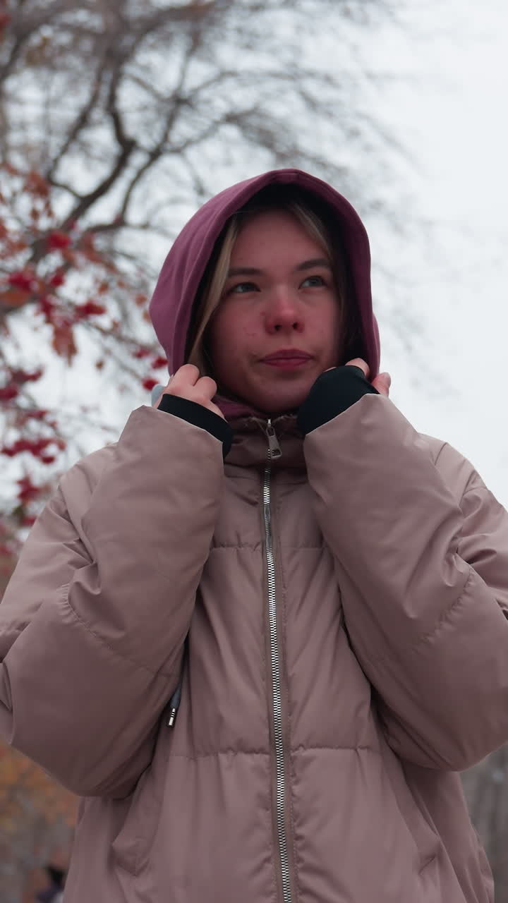 mujer envuelta en ropa de invierno cálida ajusta su sudadera con capucha mientras camina a través de un parque nevado, sostiene sus manos cerca de su cara para mantenerse caliente, rodeada de árboles desnudos con bayas rojas