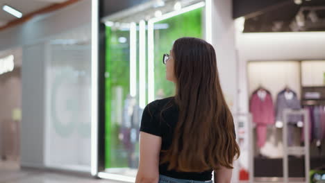 back view of lady in black top with phone in pocket walking through mall, confidently flaunting hair, with retail store displaying clothes in the background