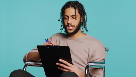 hombre en silla de ruedas escribiendo en el clipboard con bolígrafo, fondo de estudio