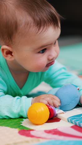 sweet little boy holds a toy in one hand and reaches for a ball with another hand. cute child lies on belly on mat playing. vertical video