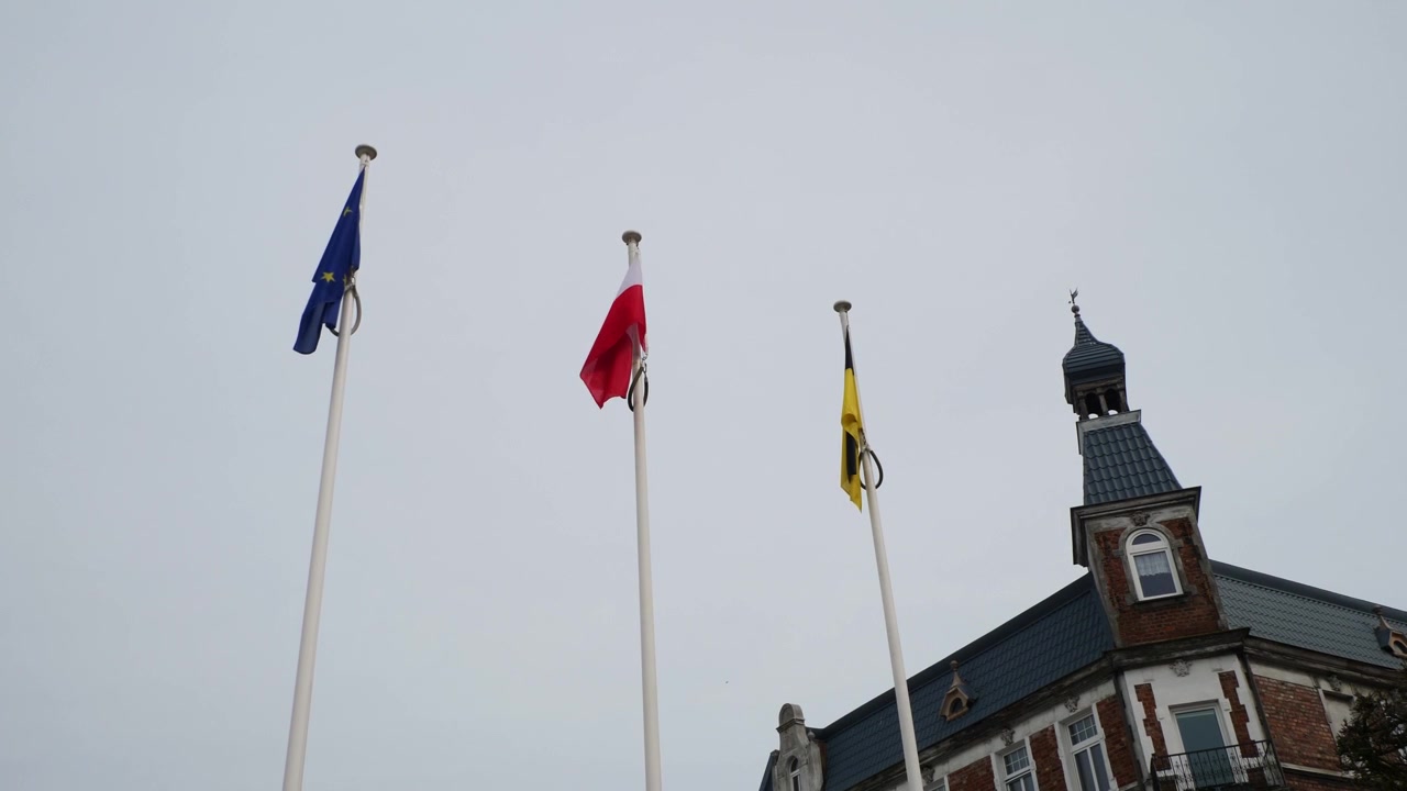 Inquadratura fissa delle bandiere comunale, nazionale e dell'UE che sventolano sulla storica piazza principale acciottolata di Puck, Polonia. Scena diurna luminosa