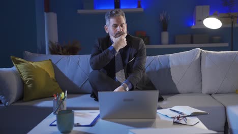 confused thoughtful businessman sitting on sofa at home and looking doubtful and serious at laptop.