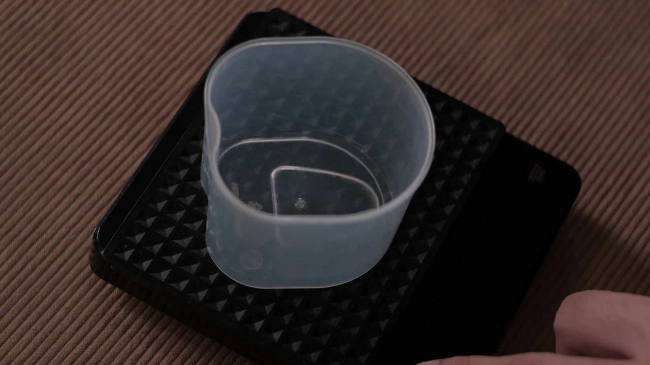 Static shot of a hand placing a plastic container on a digital scale and turning it on for indoor coffee preparation