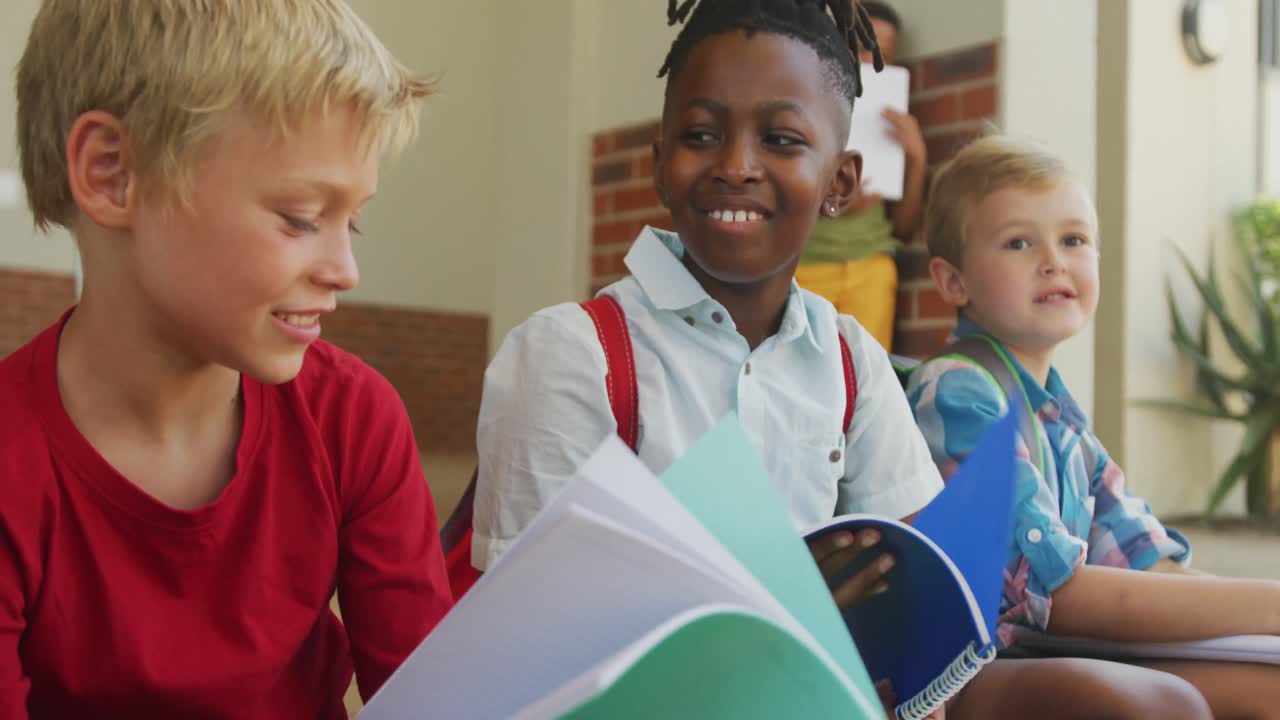 Video of happy diverse boys holding books and talking in front of school