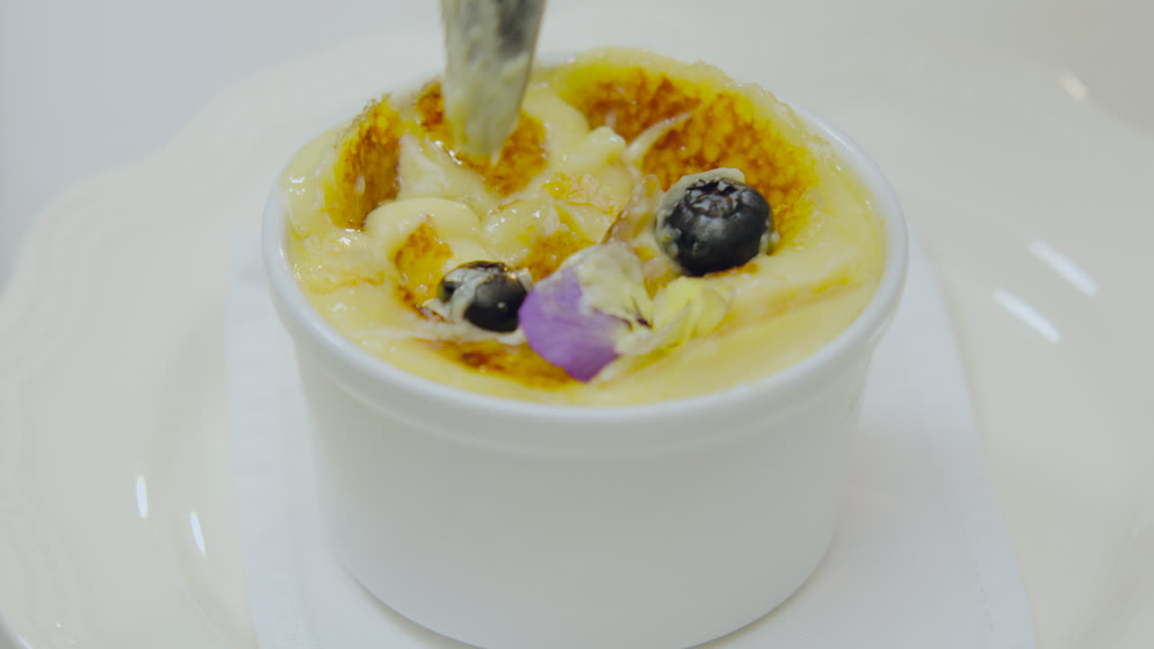 Close-up of a completely golden creme brulee, decorated with an edible flower and fresh blueberries, while a spoon cuts its caramelized surface