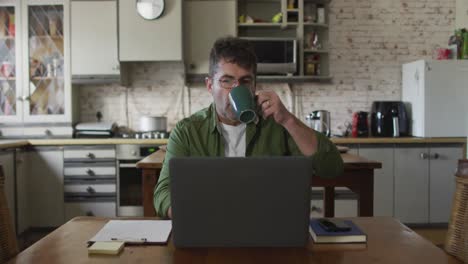 caucasian man sitting at table, using laptop and drinking coffee at home