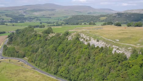 Drone-footage-moving-sideways-and-panning,-showing-a-tree-lined-steep-limestone-crag-with-fields,-dry-stone-walls,-farmland-and-the-beautiful-rural-Yorkshire,-UK-countryside-and-hills-in-the-distance