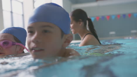 Female-Coach-In-Water-With-Children-Gives-Swimming-Lesson-In-Indoor-Pool