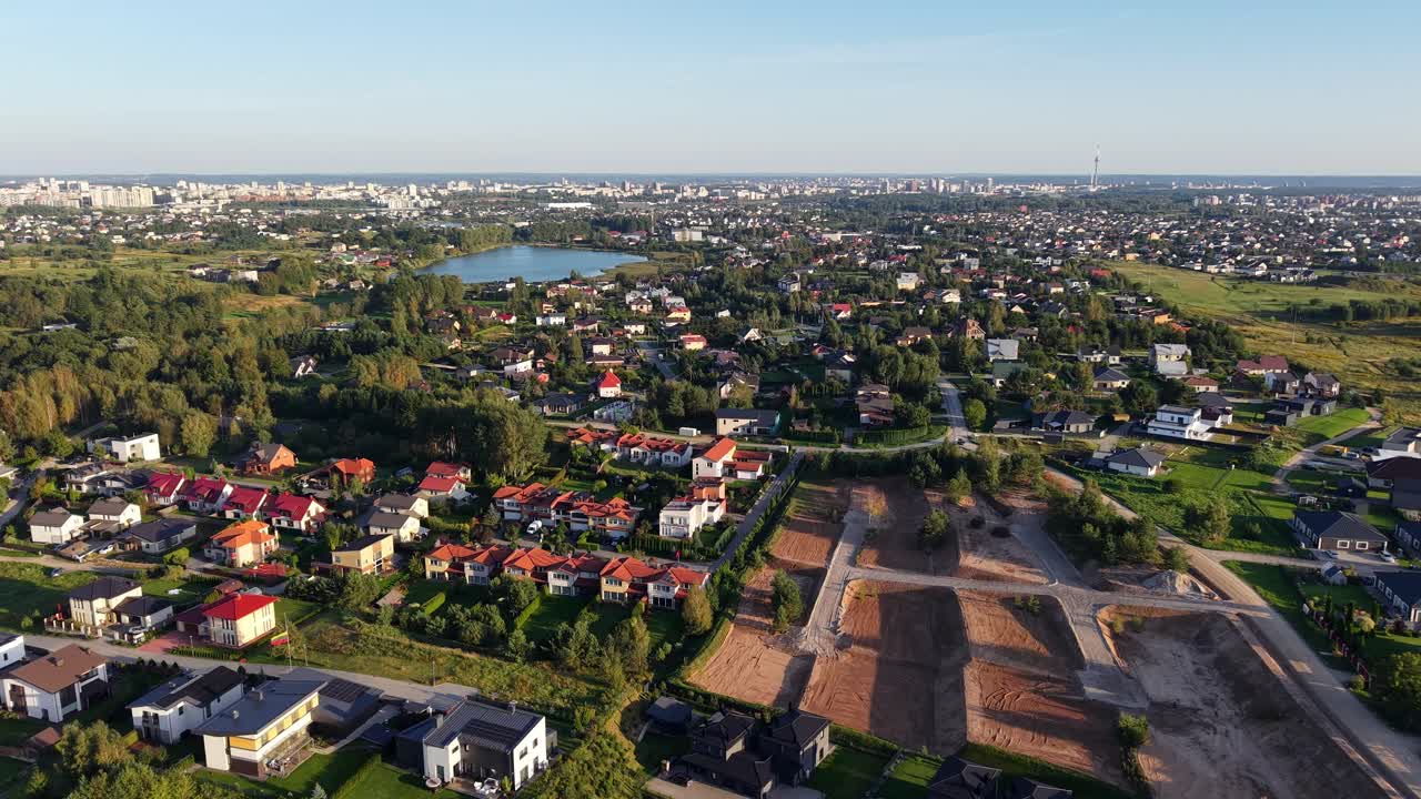 Ripresa aerea in drone sopra il villaggio di Gilužiai che mostra case, appezzamenti non edificati, uno specchio d'acqua e lo skyline della città in lontananza vicino a Vilnius, Lituania