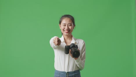 fotógrafa asiática usando una cámara tomando fotos y sonriendo tocando su pecho mientras está de pie en el fondo de la pantalla verde en el estudio