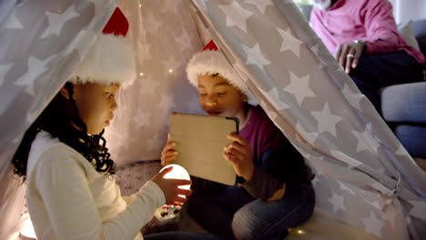 Happy-african-american-brother-and-sister-in-christmas-hats-with-tablet-in-blanket-tent,-slow-motion