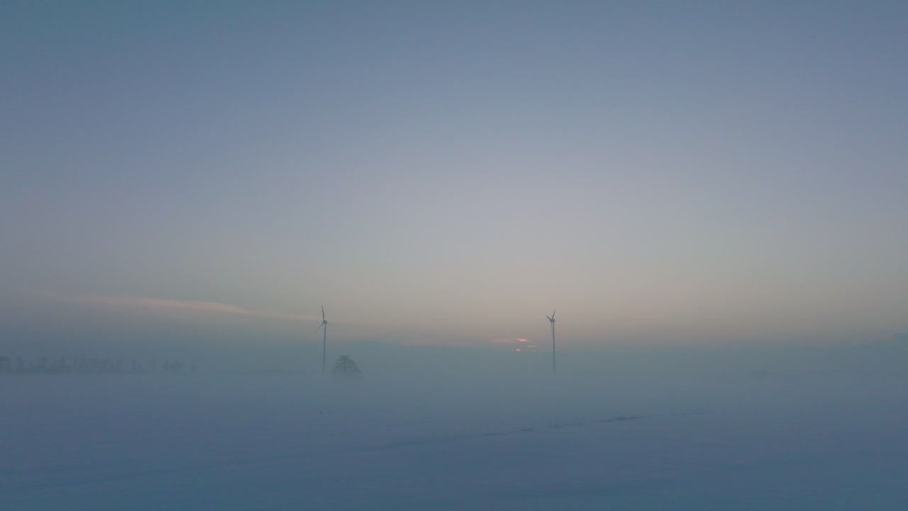 풍력 발전소에서 재생 가능한 에너지를 생성하는 풍력 터빈의 공중 전망, 안개로 가득 찬 눈으로 가득 찬 시골 풍경, 황금 시간 빛이 있는 화창한 겨울 저녁, 낮은 드론 샷이 뒤로 이동