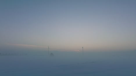 Vista-Aérea-De-Turbinas-Eólicas-Que-Generan-Energía-Renovable-En-El-Parque-Eólico,-Paisaje-Rural-Lleno-De-Nieve-Con-Niebla,-Tarde-Soleada-De-Invierno-Con-Luz-De-Hora-Dorada,-Tiro-Bajo-De-Drones-Retrocediendo