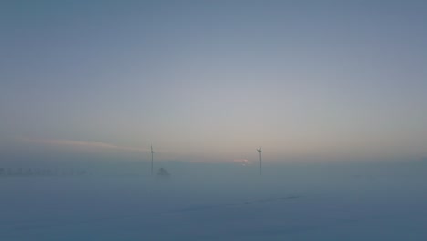 aerial view of wind turbines generating renewable energy in the wind farm, snow filled countryside landscape with fog, sunny winter evening with golden hour light, low drone shot moving backward