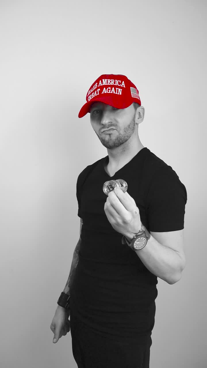 Grayscale footage of a man with tattoos and a red colored MAGA Make America Great Again Trump hat showing three Bitcoin reflective cryptocurrency coins and nodding with approval. Man is happy.