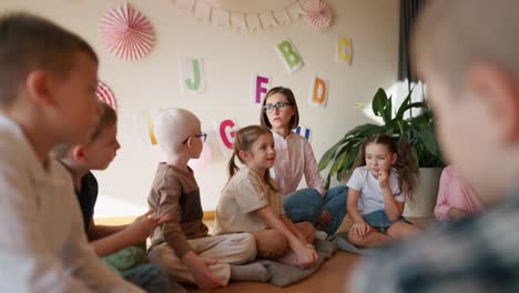 una chica rubia con un peinado bob en gafas y una camisa blanca se comunica con un niño albino en gafas durante una lección en un club para preparar a los niños para la escuela