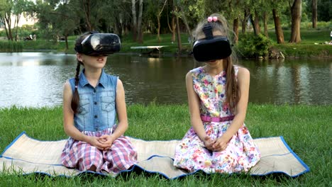 two little girls explore the surrounding space in virtual reality helmets. they sit on the lawn by the river waving and laughing.