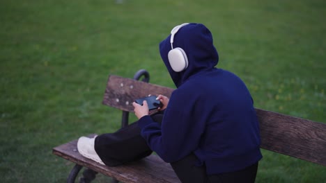 Mujer-Solitaria-Con-Capucha-Y-Auriculares-Sentada-En-Un-Banco-De-Madera-Y-Usando-Un-Teléfono-Inteligente